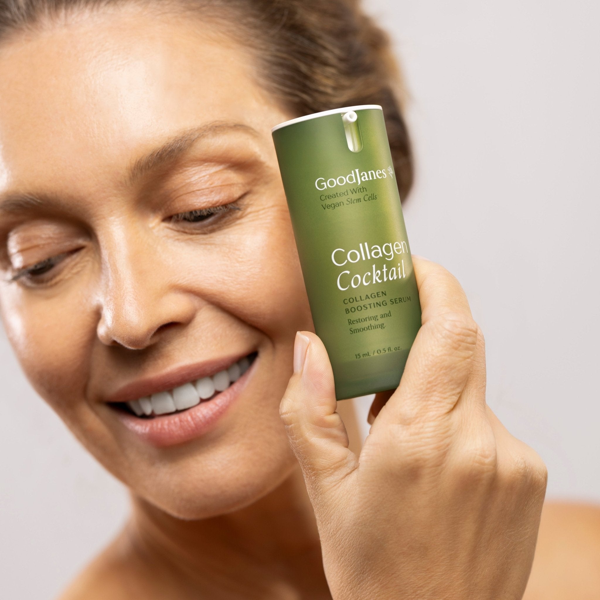 Woman applying cream to her face with a bottle of Goodfellow's Collagen Cocktail in front of a gray background