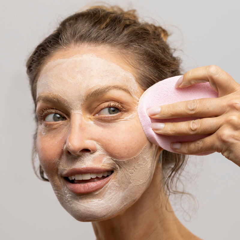 Woman applying a facial mask with a pink brush on a plain background
